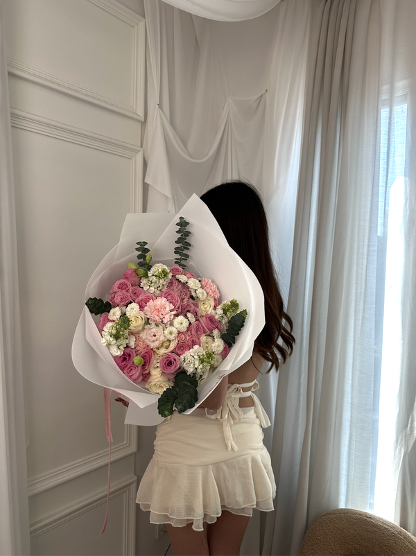 A woman stands with her back to the camera holding a large bouquet of pink, white, and cream roses wrapped in white paper, set against a soft white curtain and paneled wall.