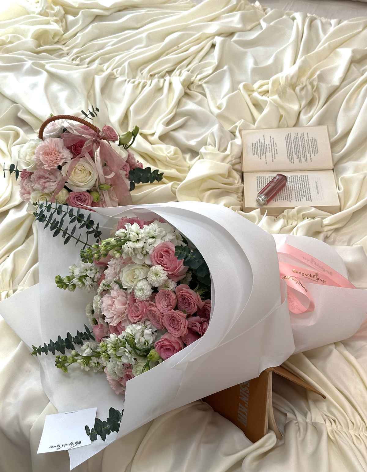 A floral bouquet wrapped in white paper with pink and white flowers and green foliage, placed on a cream satin fabric surface beside two books and a small bottle.