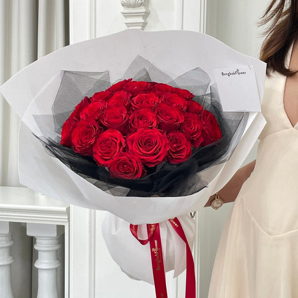 Bouquet of red roses wrapped in black paper with a red ribbon, held by a person in a white dress.