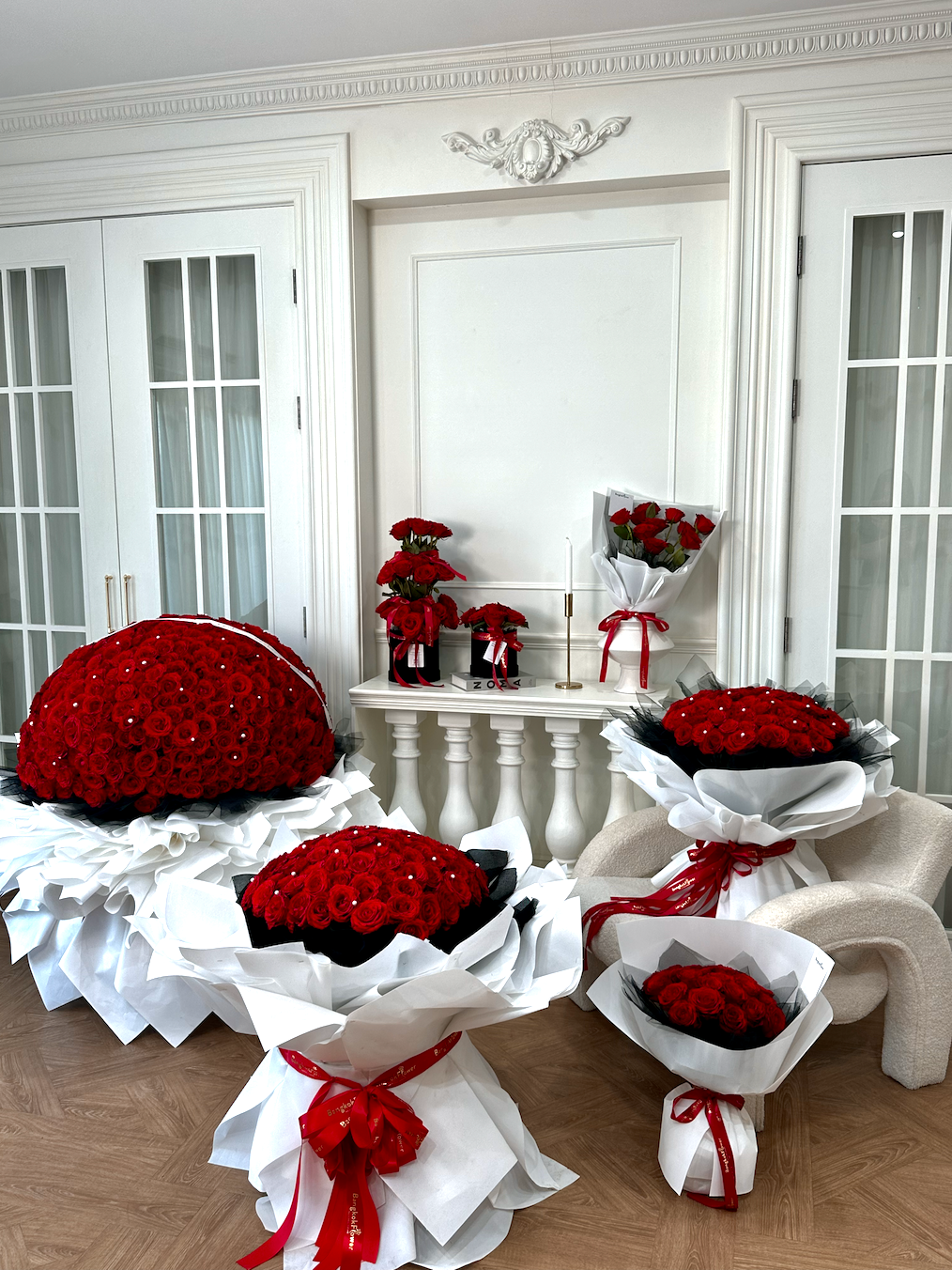 Multiple large and small arrangements of red roses wrapped in white paper with red ribbon bows, displayed on a chair and table in a bright white room with French doors.