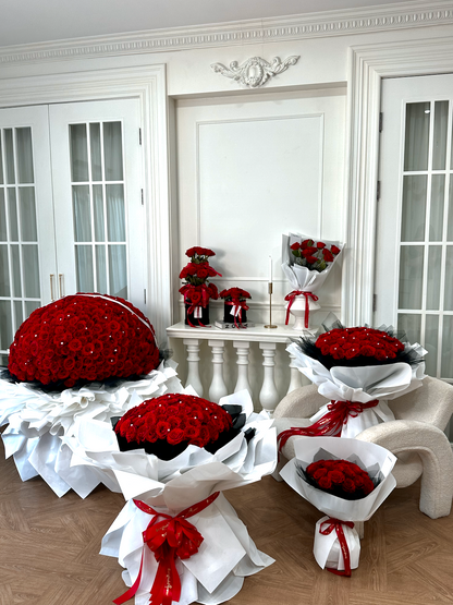 Multiple large and small arrangements of red roses wrapped in white paper with red ribbon bows, displayed on a chair and table in a bright white room with French doors.
