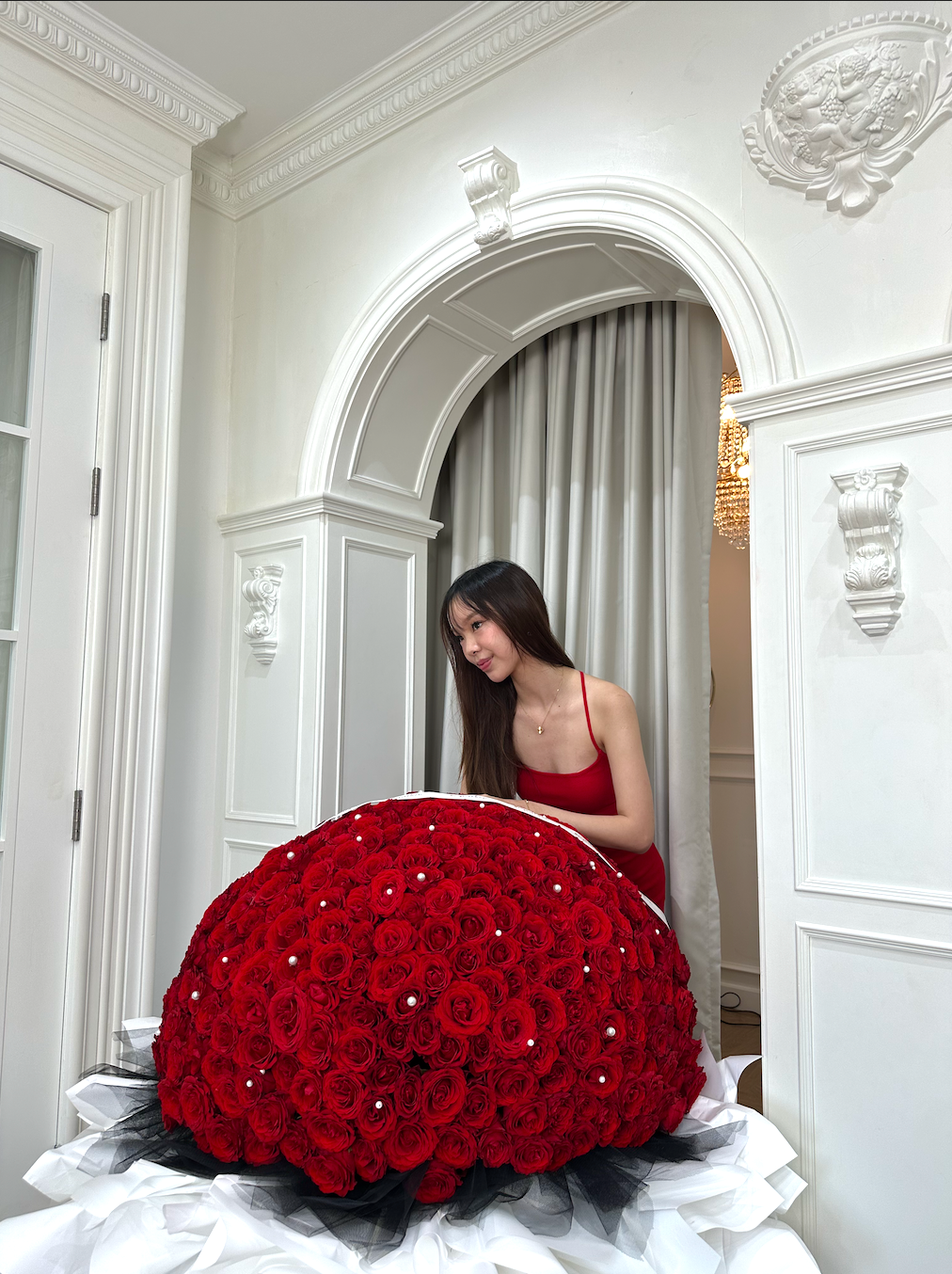 A large, spherical bouquet of deep red roses with a black tulle base and white ribbon, displayed on a table in an elegant white room with an arched doorway behind where a woman in a red dress is seated.