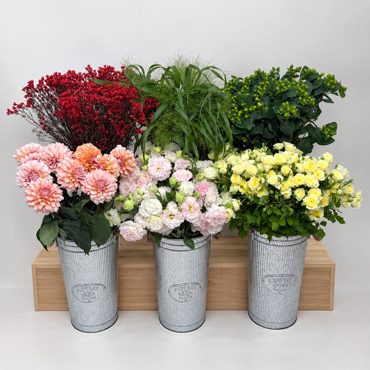 Three galvanized metal flower baskets filled with assorted faux flowers and greenery, arranged on a wooden riser against a light gray background.