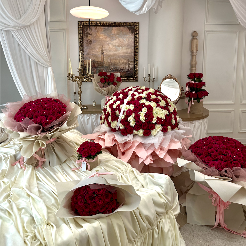 Multiple large bouquets of red and white roses arranged on draped white fabric in an elegant indoor room with white drapery, a framed painting, and candle holders in the background.