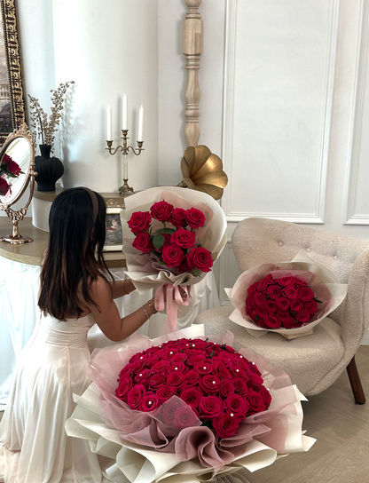 A woman in a white dress stands beside three large arrangements of red rose bouquets wrapped in white and blush pink paper, set in a bright, elegant room with a beige armchair and decorative wall details.