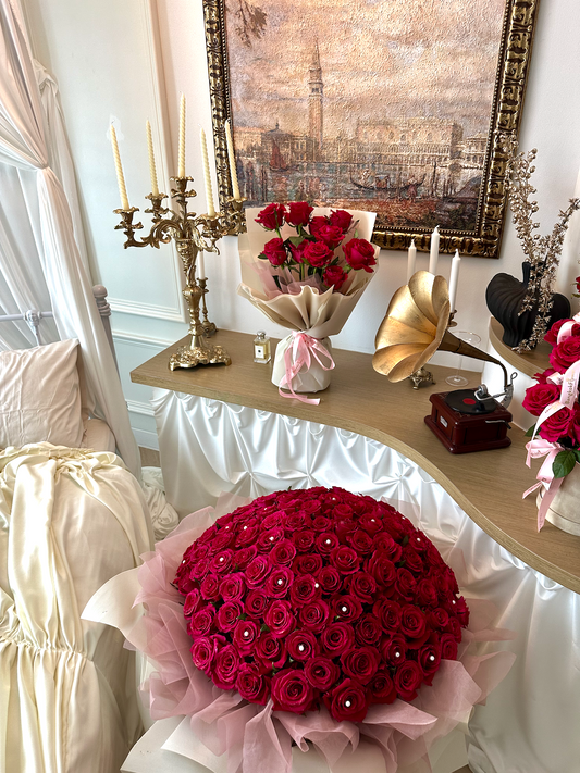 A large round bouquet of deep red roses with small pearl-like center accents and pink tulle wrapping, displayed in a decorated room setting on a console table with candles, a gramophone, and framed wall art.